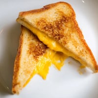 Golden-brown grilled cheese sandwich with gooey melted cheddar on buttery sourdough, cut diagonally and served beside a steaming bowl of tomato soup.